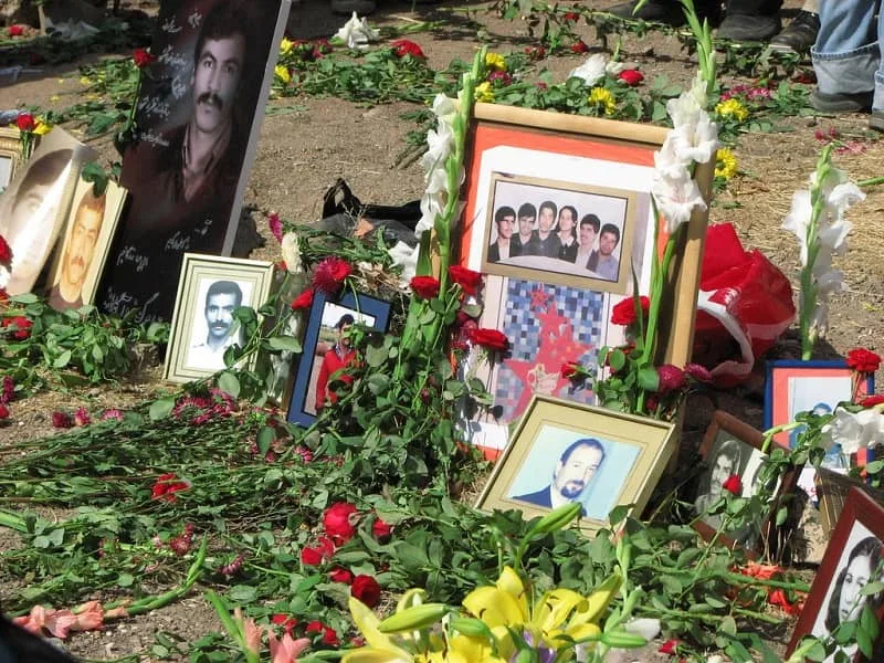 Framed portraits of men and a group photograph, surrounded by carnations and white gladioli on bare ground.