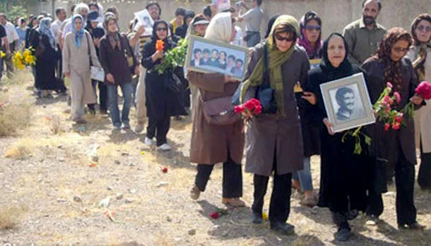 A line of women walking along a dirt path, several carrying framed portraits and flowers.